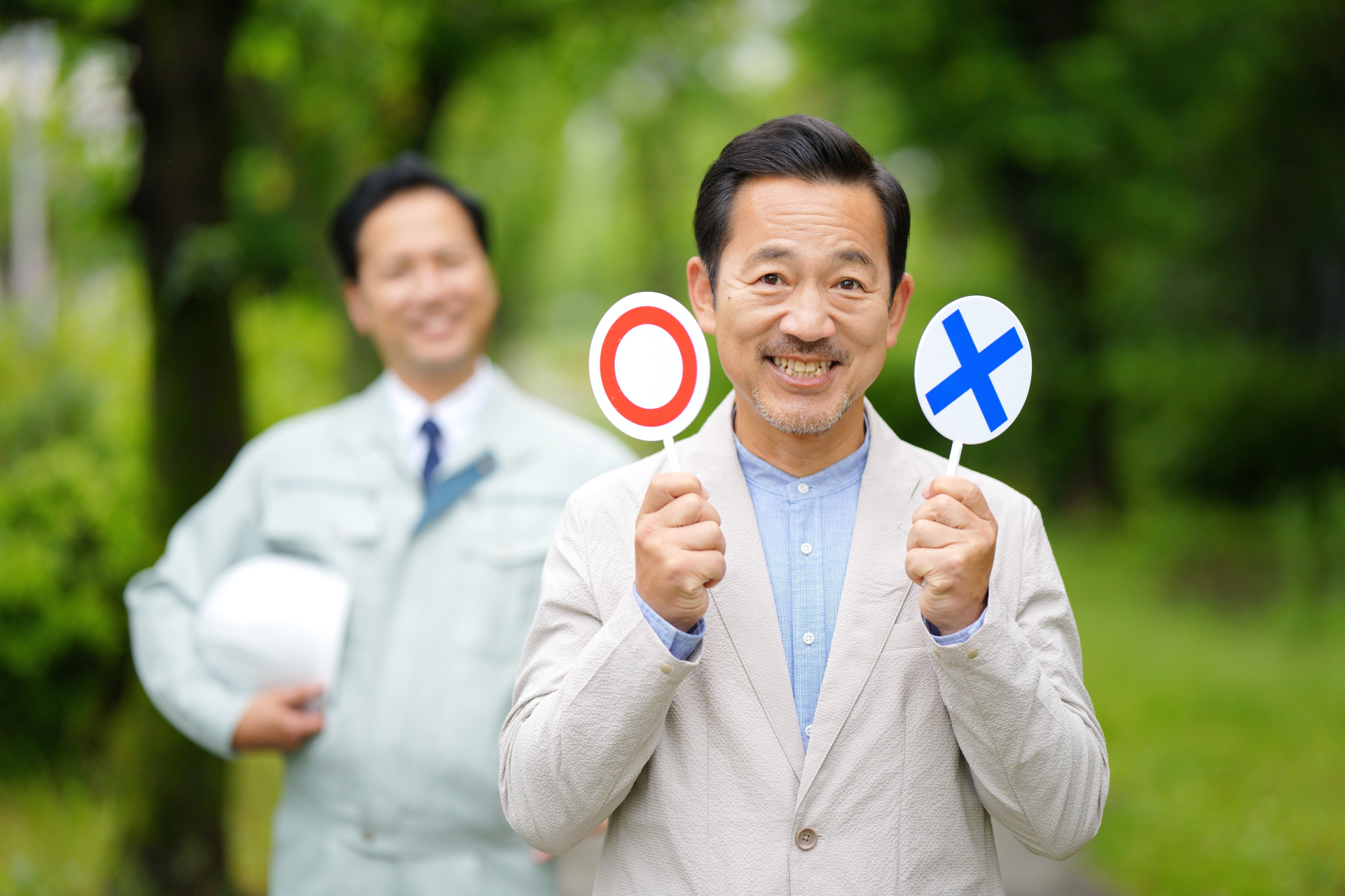 公園でマルバツの札を持つ中年男性と作業服の男性