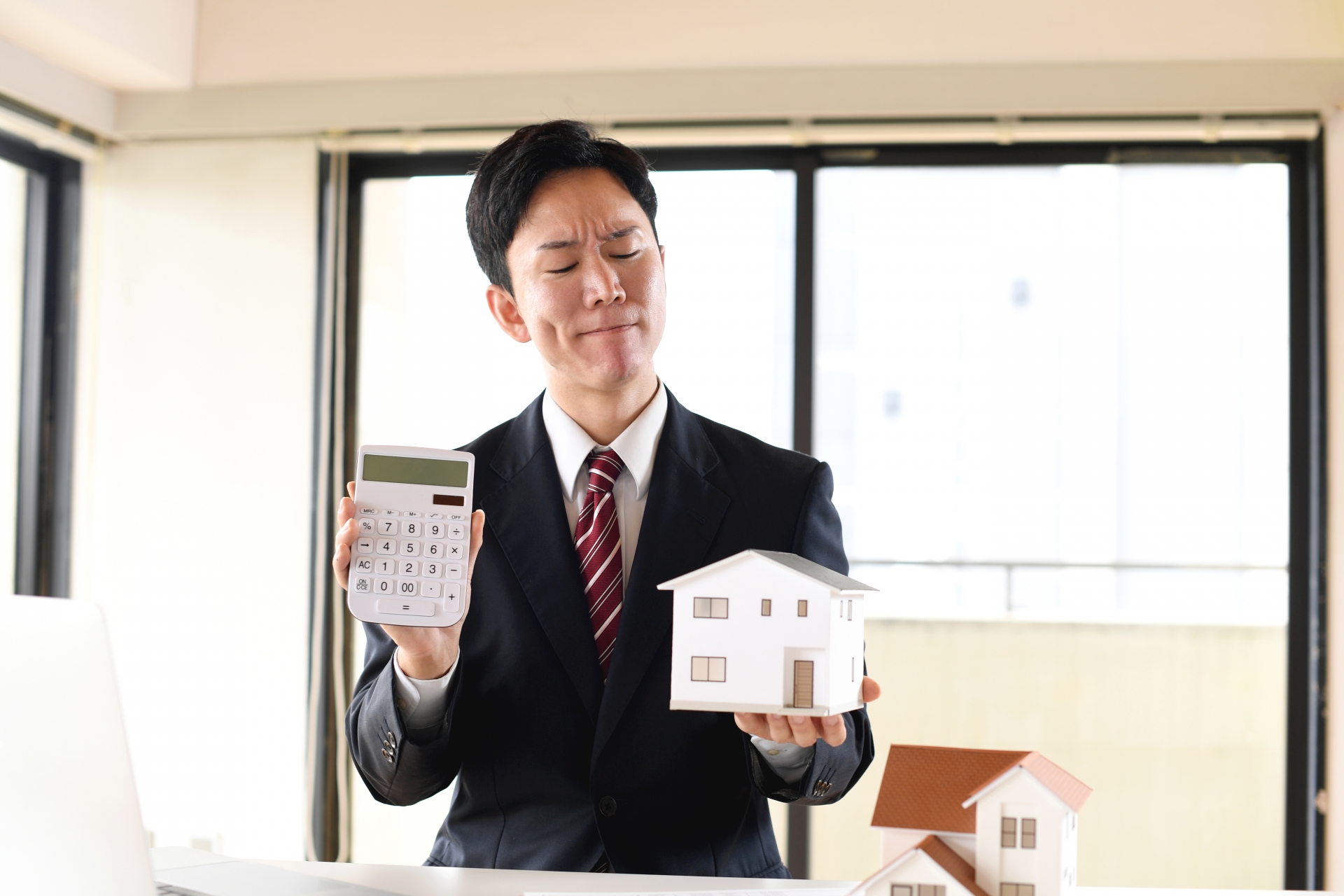 住宅模型と電卓を持ち悩むスーツ姿の男性